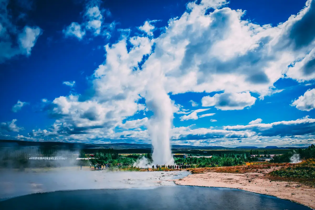 Visitors standing near Strokkur geyser as it erupts high into the air under a clear summer sky