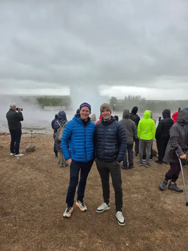 Two Golden Circle Iceland team members standing in front of Strokkur geyser erupting at Geysir